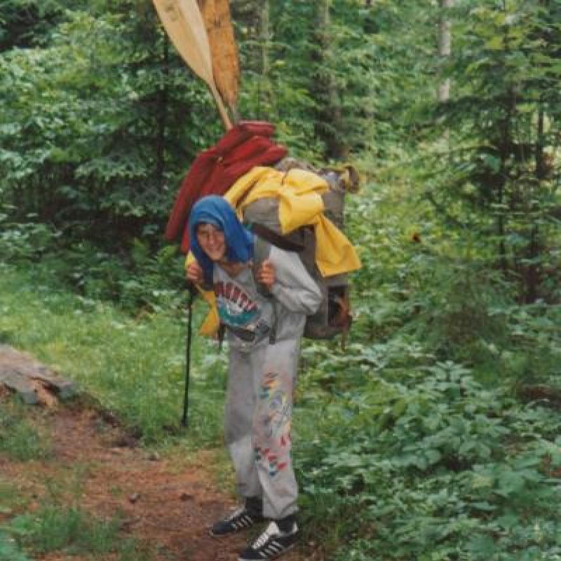 A camper struggles under the weight of a large backpack. Two canoe oars protrude from the sack.