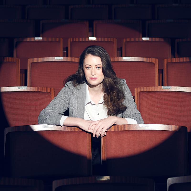 Woman sitting in an empty theatre, looking straight ahead, with a spotlight on her.