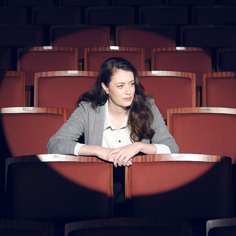 Woman sitting in an empty theatre, looking to the side, with a spotlight on her.