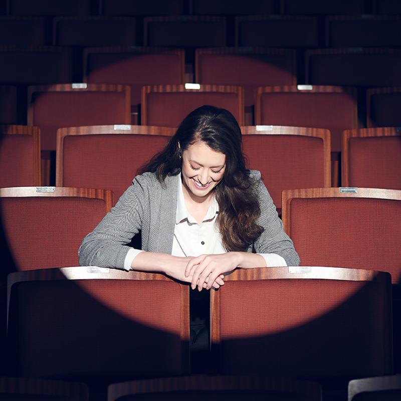 Woman sitting in an empty theatre, looking down and laughing, with a spotlight on her.