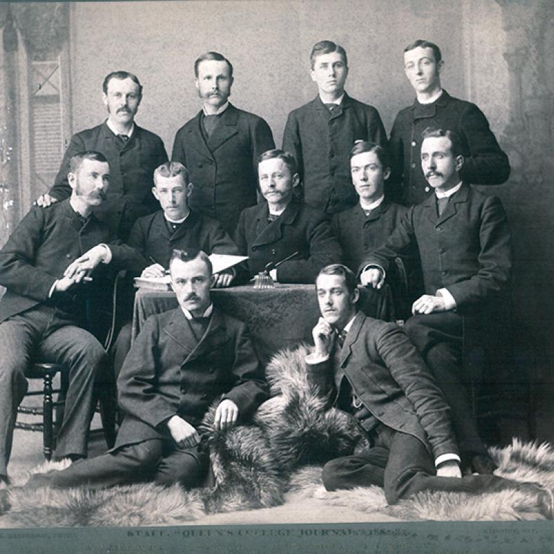 Members of the Queen's Journal in 1883. Nine men sit around a table with two men sitting in front of the table on fur rugs..