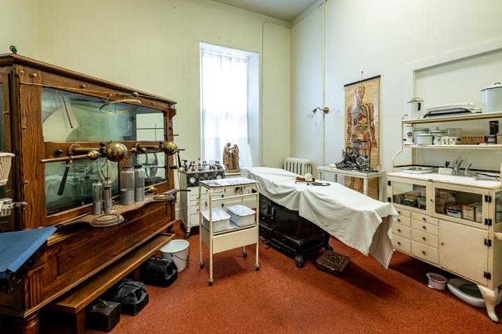 A full room view of various medical equipment, including an examination table, a full-size chart that shows the organs in a human body. 
