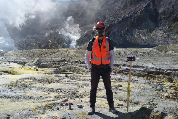 David McLagan standing on an active volcano wearing a hardhat, safety mask and orange vest.