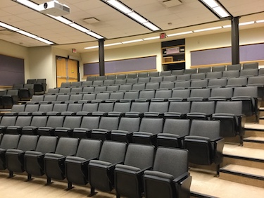 Tiered classroom with grey auditorium seats, wood doors, and purple accents on the wall.
