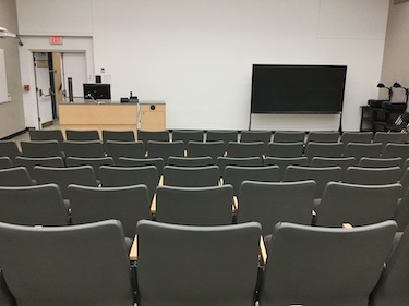 Student view in a large auditorium with grey chairs and a white board and blackboard at the front of the room.
