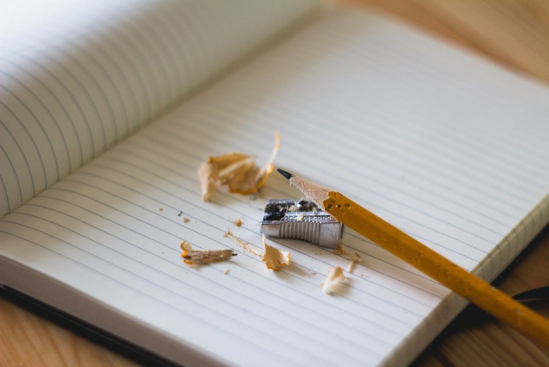 "Pencil leaning on an open lined notebook with a small metal pencil sharpener and pencil shards beside it"