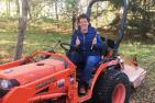 Classics professor Barbara Reeves holds excavating tools as she sits on an orange Kubota tractor.
