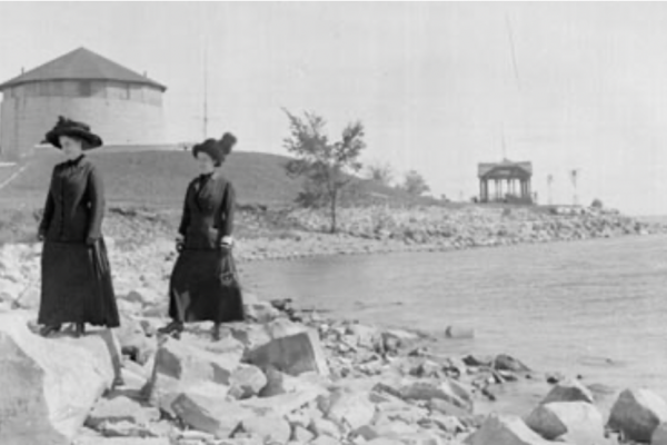 A black and white photograph of two women standing on the rocks at Macdonald Park on the shore of Lake Ontario with Murney Tower in the background.