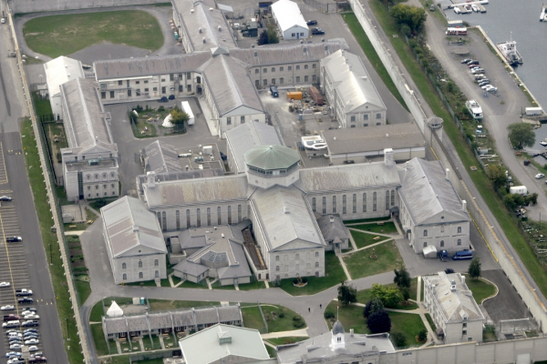 An aerial image of Kingston Penitentiary where you can see the stone entryway, two central "t" shaped buildings, and a number of various outbuildings with a parking lot on the left 