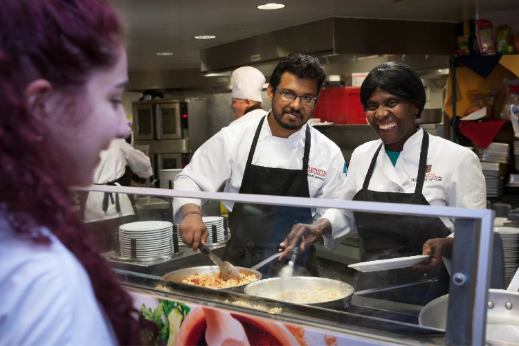 [Queen’s Hospitality Services staff members serve a student in a Queen’s dining hall.]