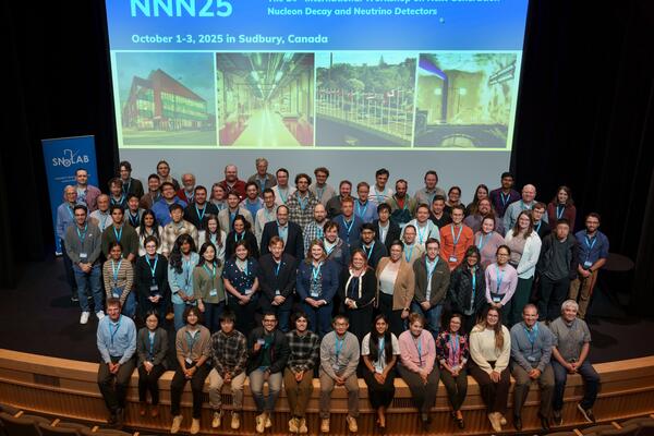 A large group of people sit on a stage. A logo is projected behind them on a screen.