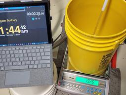A tablet with a wall clock and a stopwatch, a scale, a pail, and chiller fluid flowing into the pail