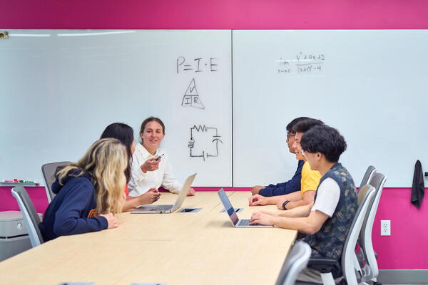 A group of students sit in front of a whiteboard with equations on it
