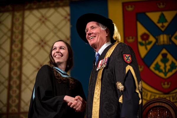 A professor and dean shake hands at convocation. They are adorned in traditional robes.
