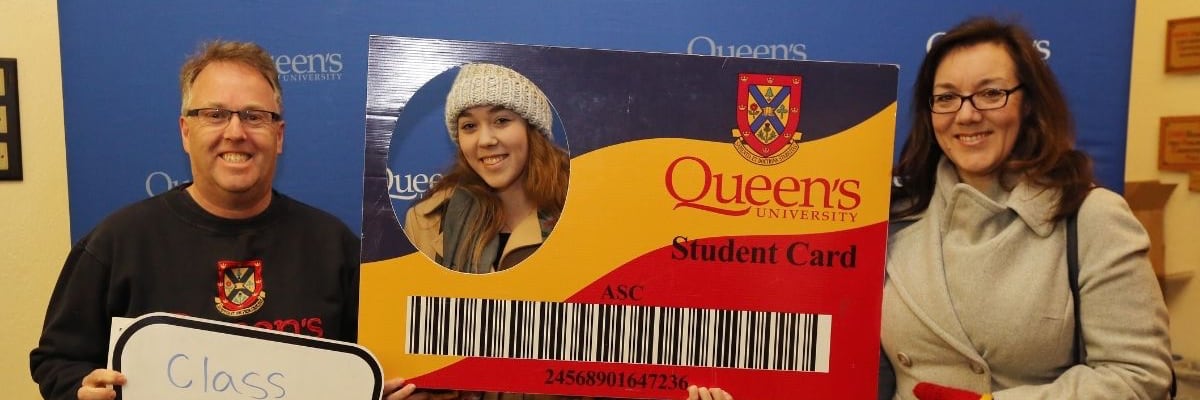 Two excited parents stand beside a smiling student holding a mockup ID card labeled "Queens Student Card."
