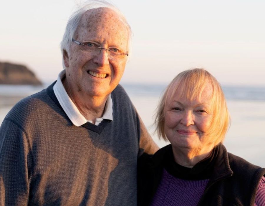 A senior couple stands on the sandy beach, overlooking the ocean under a clear sky.