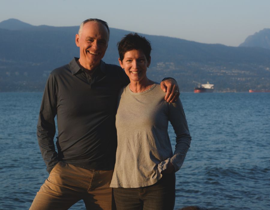 A couple smiling, close together during sunset, with a nature background, with a lake and mountains.