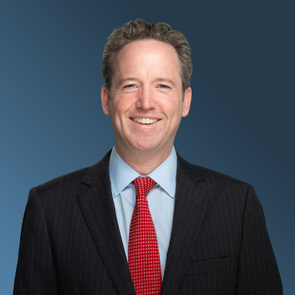 Matthew McGrath, Com’96, smiling at the camera, wearing a suit with a dark blue background.