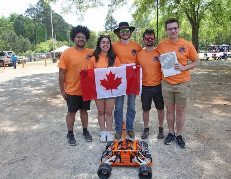 Four members of the Queen's Lunar Exploration Team in orange shirts pose with a Canadian flag at the 2025 rover challenge event.