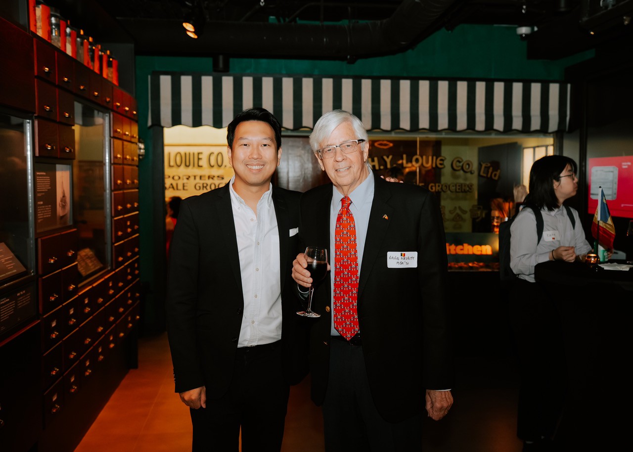 Jonathan Kong and David Nesbitt in suits, standing together at a Vancouver alumni event in a museum setting.