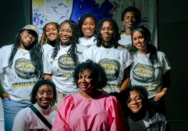 A group of people posing for a photo indoors, smiling and standing together against a wall. They are dressed casually in jeans and exhibit a sense of friendship and happiness.