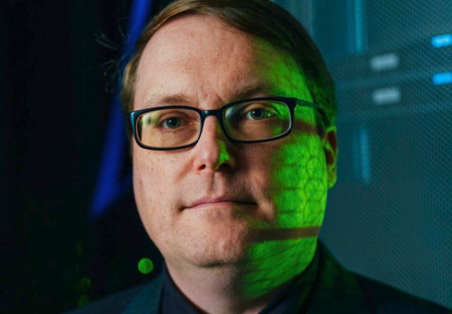 Ryan Grant, wearing glasses and a suit, stands before a computer, highlighting his contributions to supercomputing advancements.