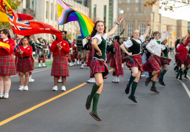 A group of Queen's students in colorful kilts joyfully dancing together on a lively street during a Homecoming celebration.