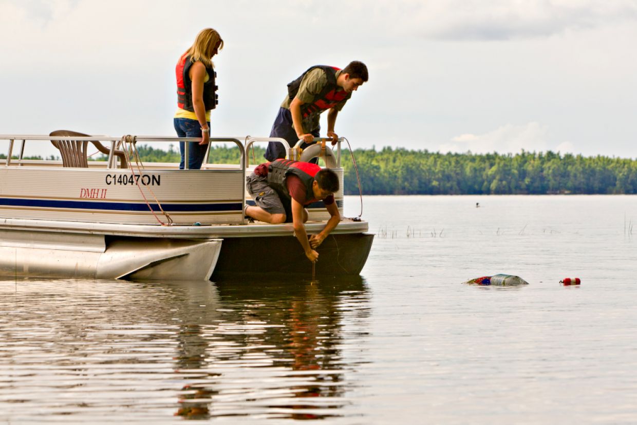 Two people wearing red life vests on a boat stand, while a third person in a red life vests kneels on the boat holding a rope that leads into the water. 