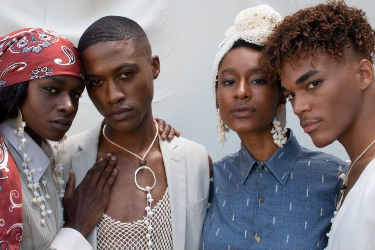 Four Black people with somber expressions stand together linking arms wearing cotton clothes.