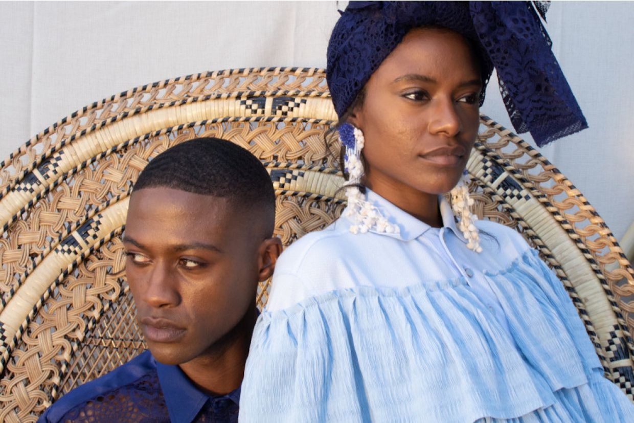 A Black man sits in a blue t-shirt with a serious expression. A Black woman in a light blue blouse, white earrings, and a dark blue hair scarf sits on his lap looking in the opposite direction. They are both on a wicker chair.