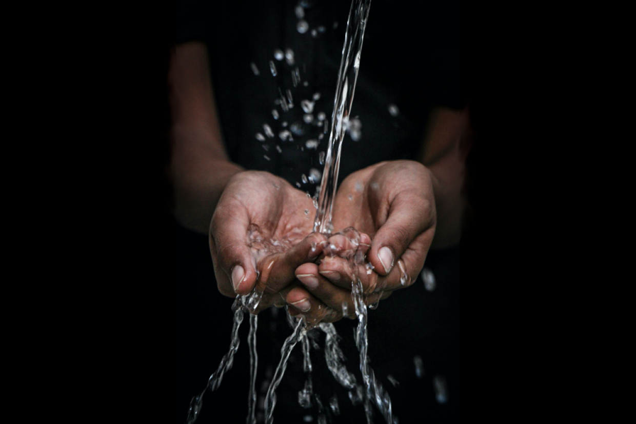 An known person has their hands outstretched and cupped. Water is poured into the hands. Some water runs between the fingers.