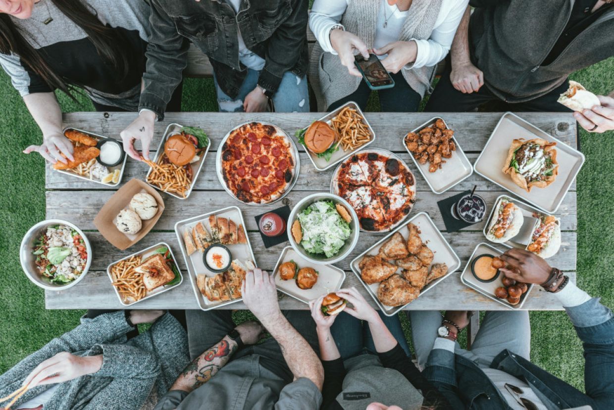 A diverse group of people gather around a table spread with pub food. 