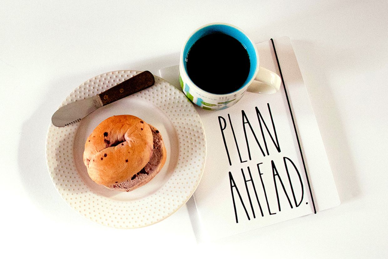 A sliced bagel on a plate with a spreading knife, coffee, and a journal titled "Plan Ahead."