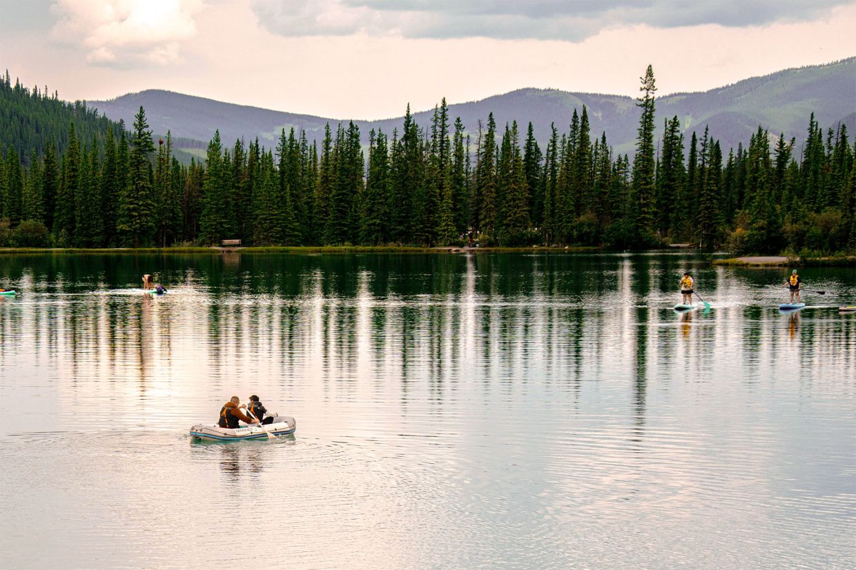 A photo from Bragg Creek with a view of a lake with people canoeing and swimming, trees, and the Rocky Mountains