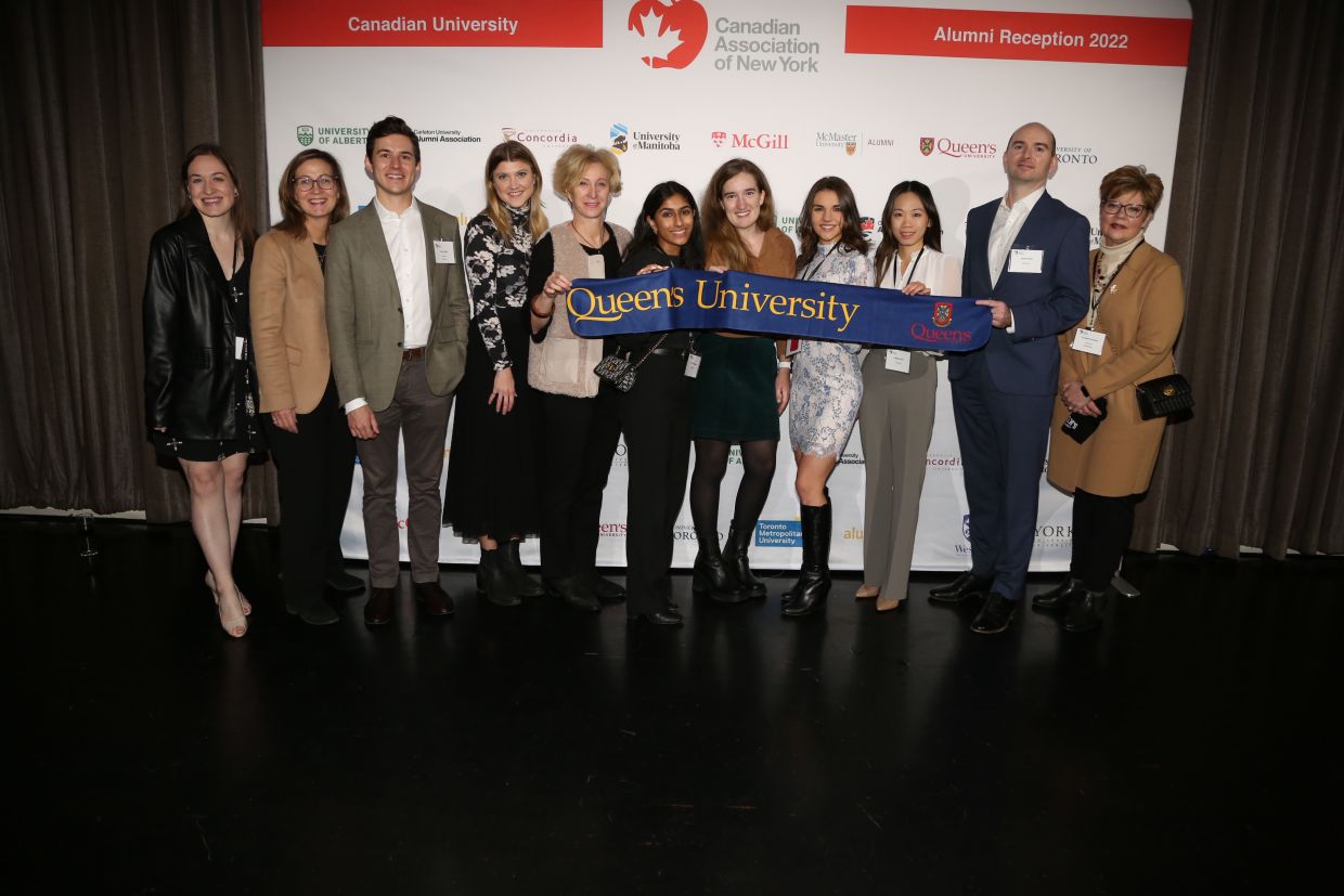 Alumni posing in front of a backdrop holding a Queen's University flag.