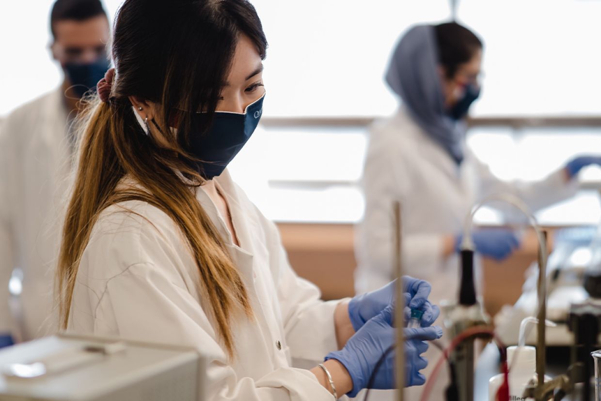A student in a lab coat, gloves, and mask works with a test tube.