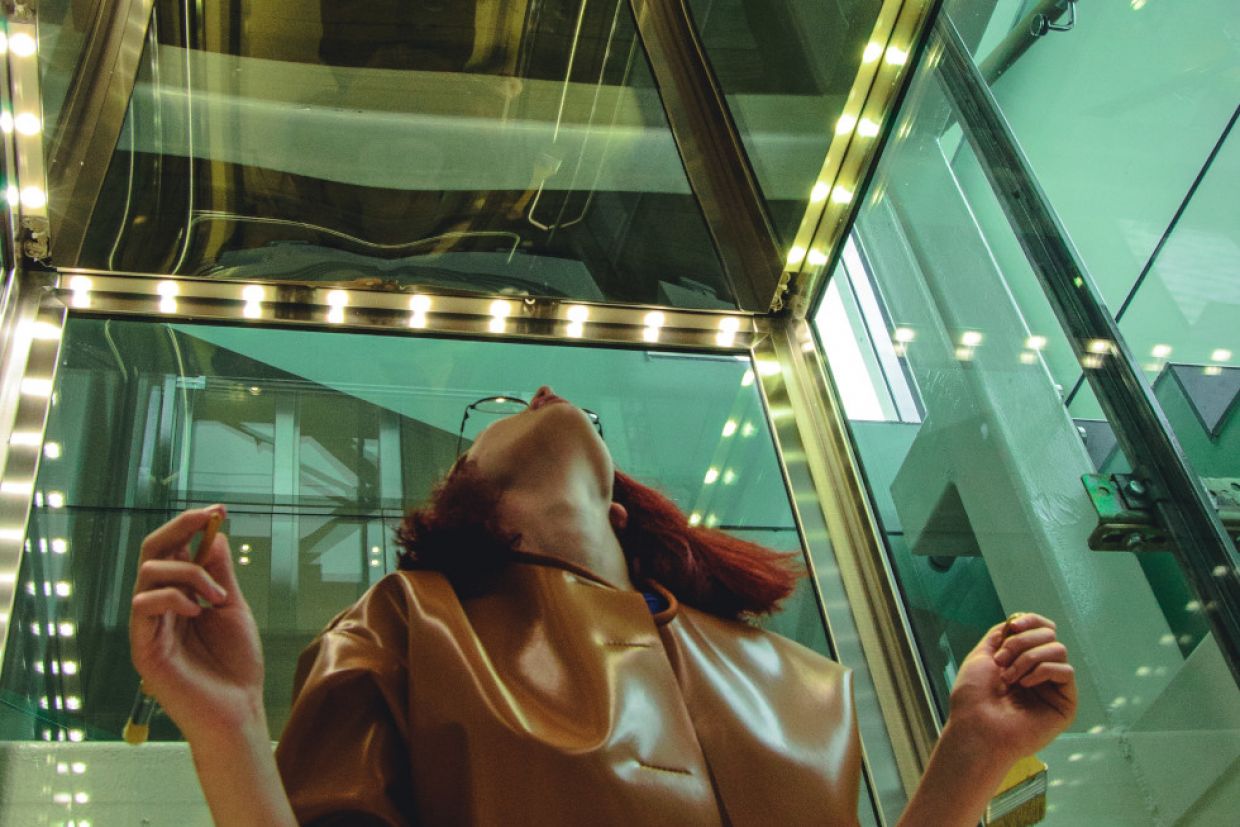 A woman in a room of mirrors, looks at a ceiling mirror perplexed.