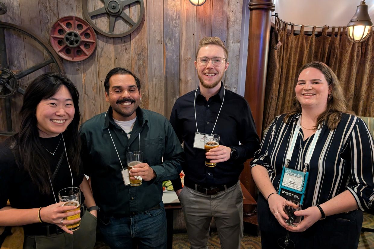 Four happy alumni gathered at Steamworks Brewpub, during the Roundup last year, standing close and holding drinks while smiling at the camera.