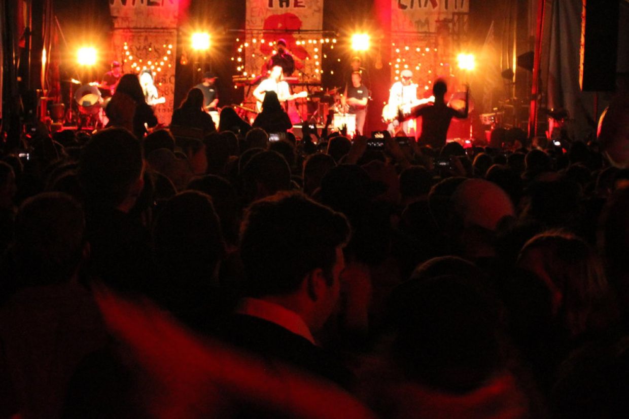 A band performs on a stage. A crowd of people cheer them on in the foreground. 