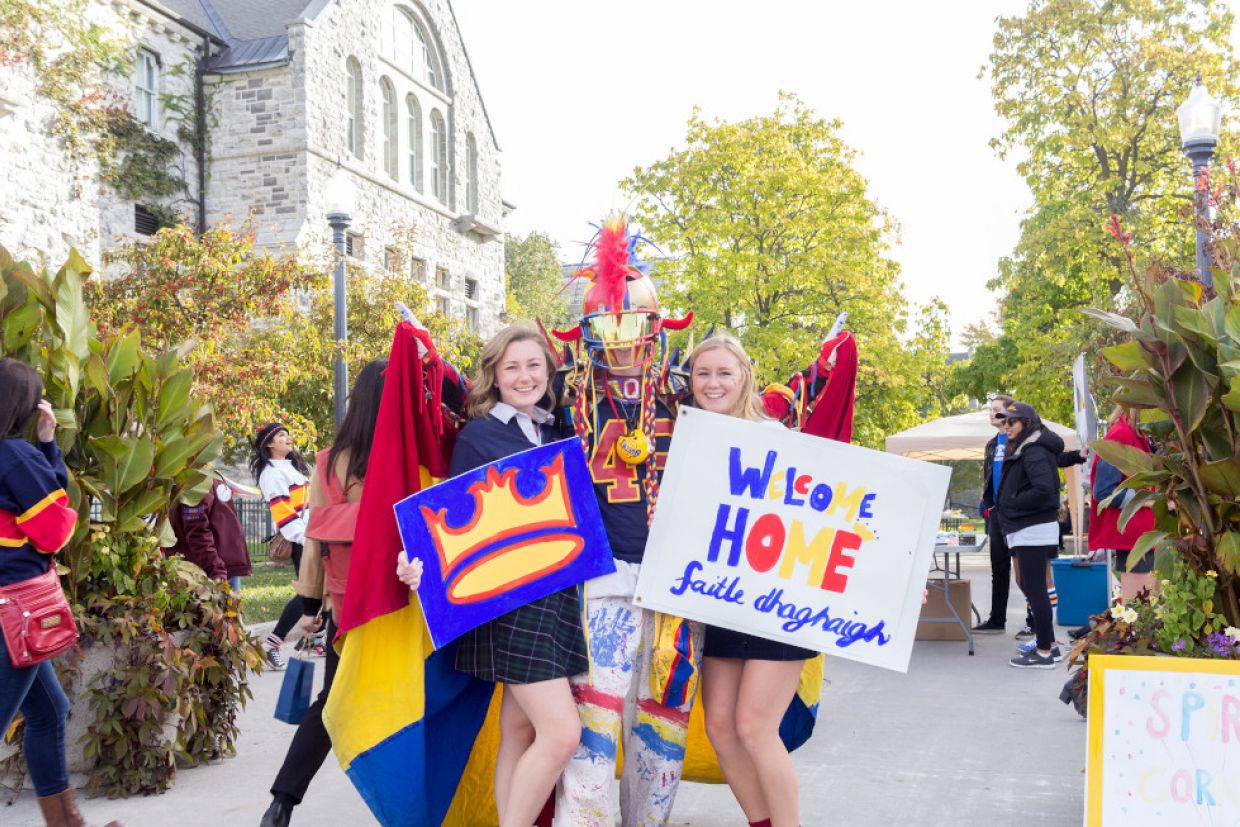 The superfan in tricolour poses for a photo with two students holding signs. One sign is the Queen's crown. Another says "welcome home."