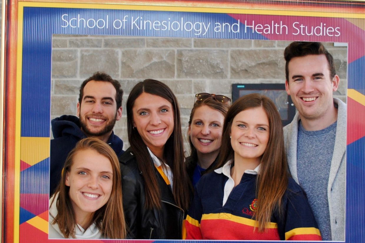 Students in tricolour clothing smile while holding a cardboard frame that says "School of Kinesiology and Health Studies"
