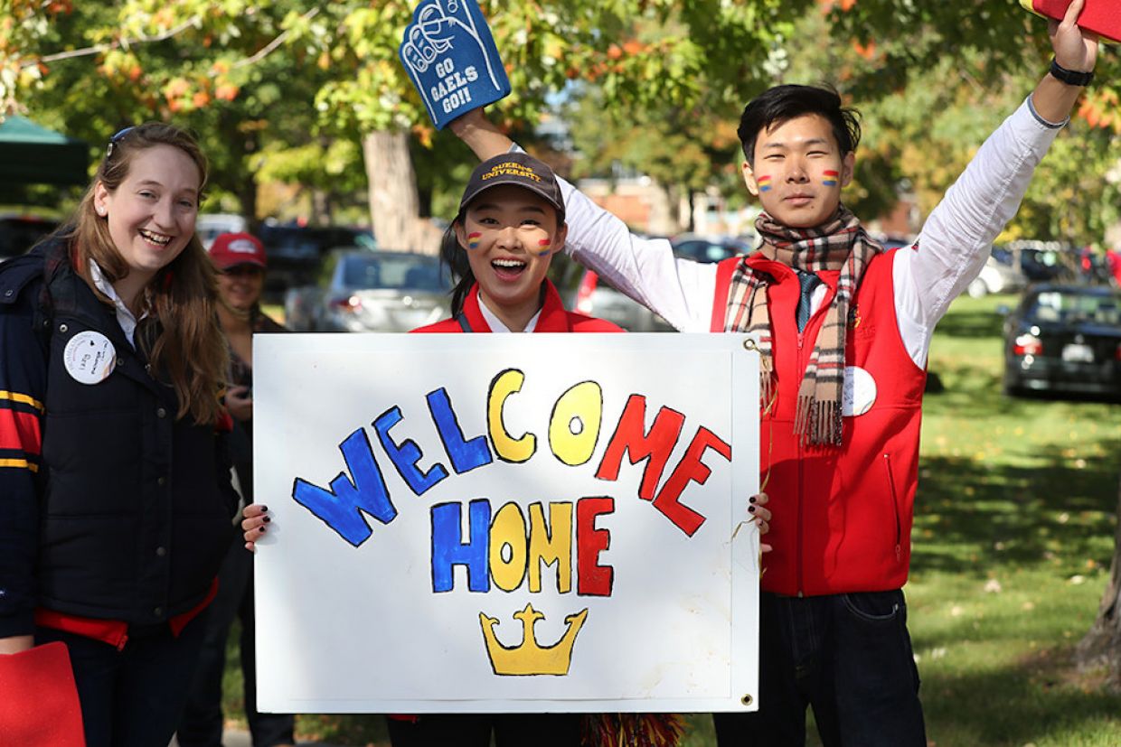 Students in tricolour welcome back alumni. One holds a sign that says "welcome home," another wears foam fingers.