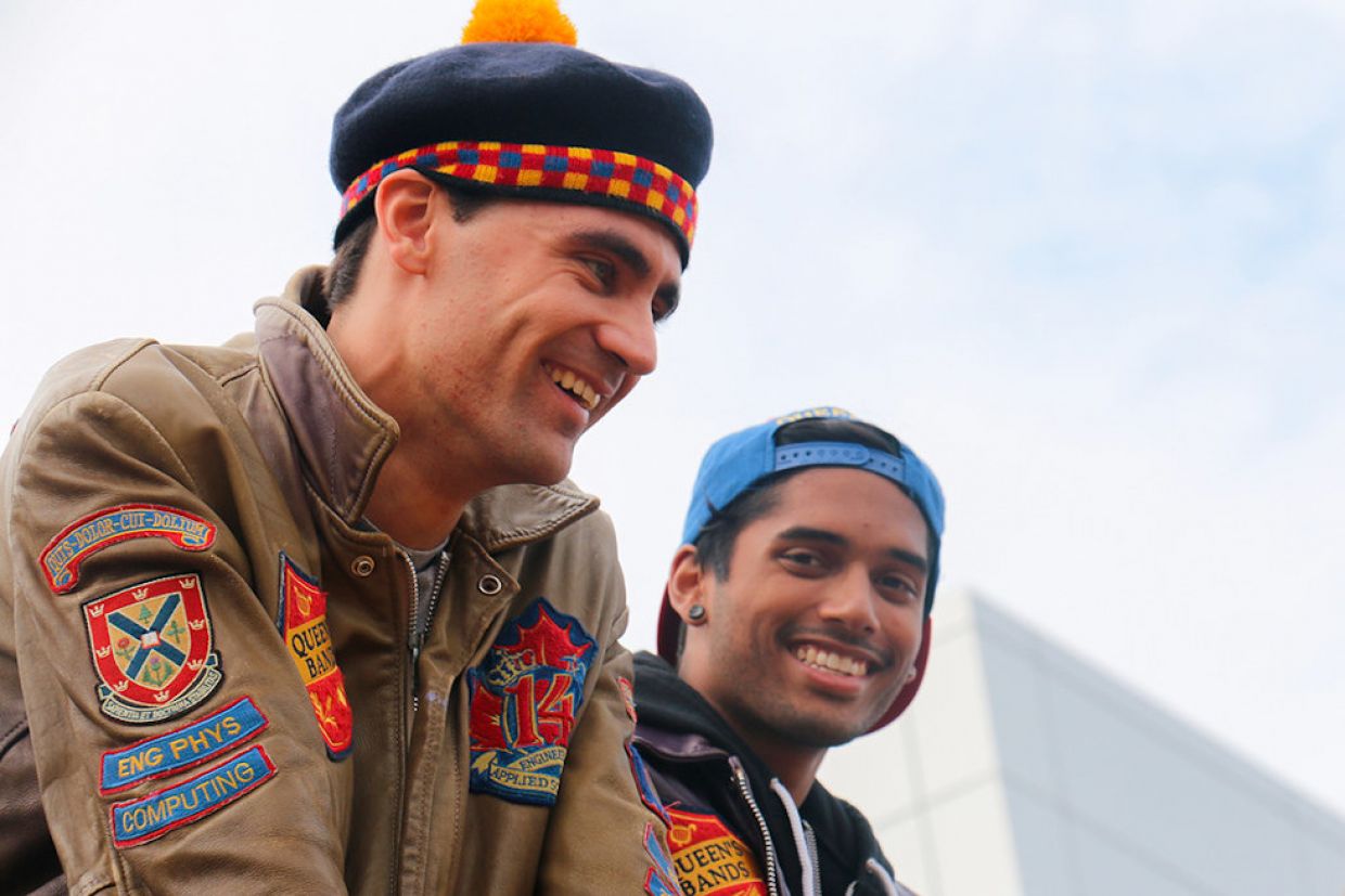 Two students watch the football game. One wears a gold engineering jacket and tam, the other wears a purple engineering jacket and baseball cap.