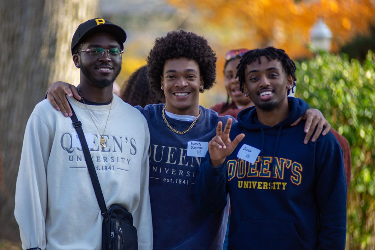 Three alumni smiling, one waiving, while wearing Queen's sweaters in autumn