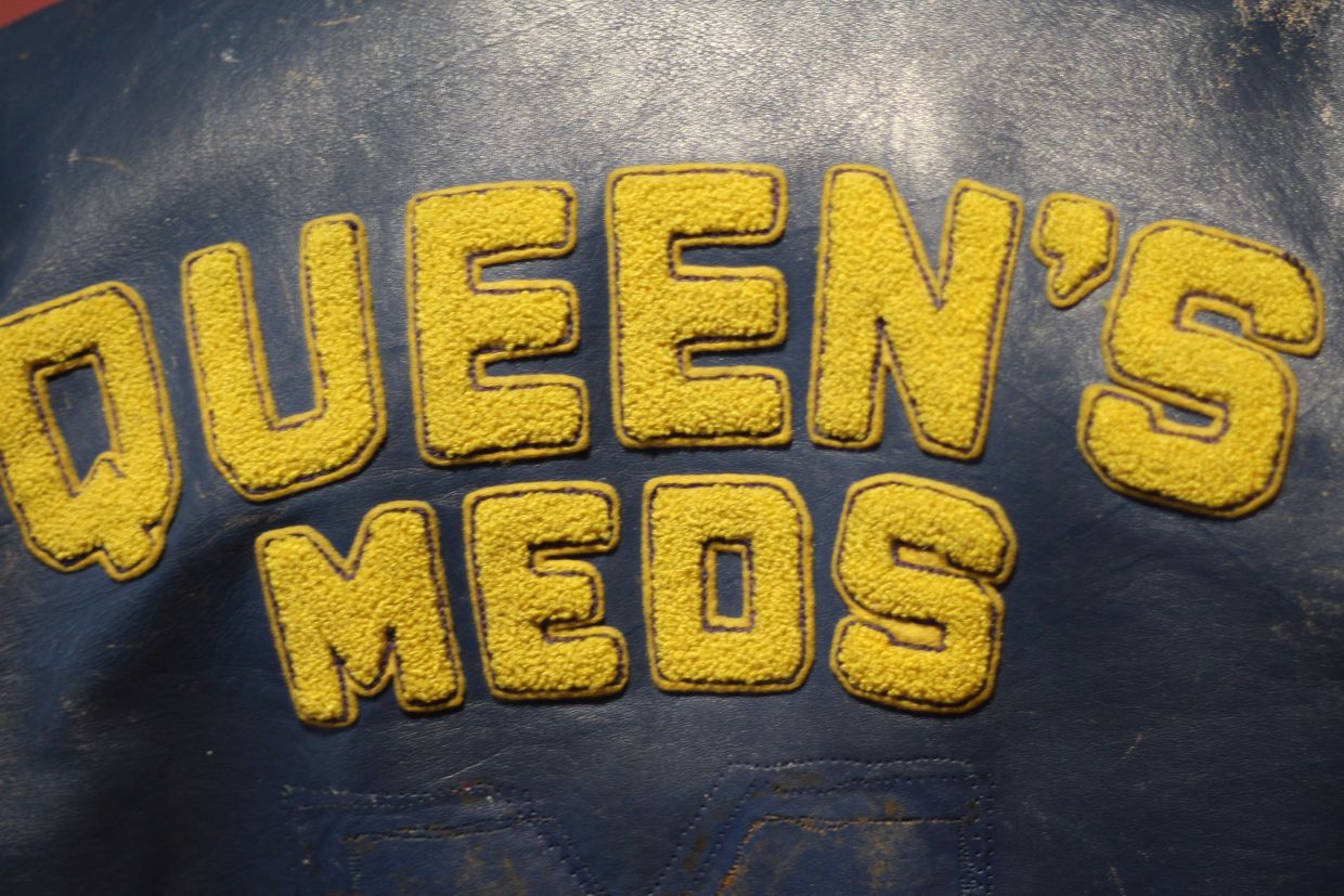 Close up of a blue leather jacket with the words "Queen's Meds" across the back in gold