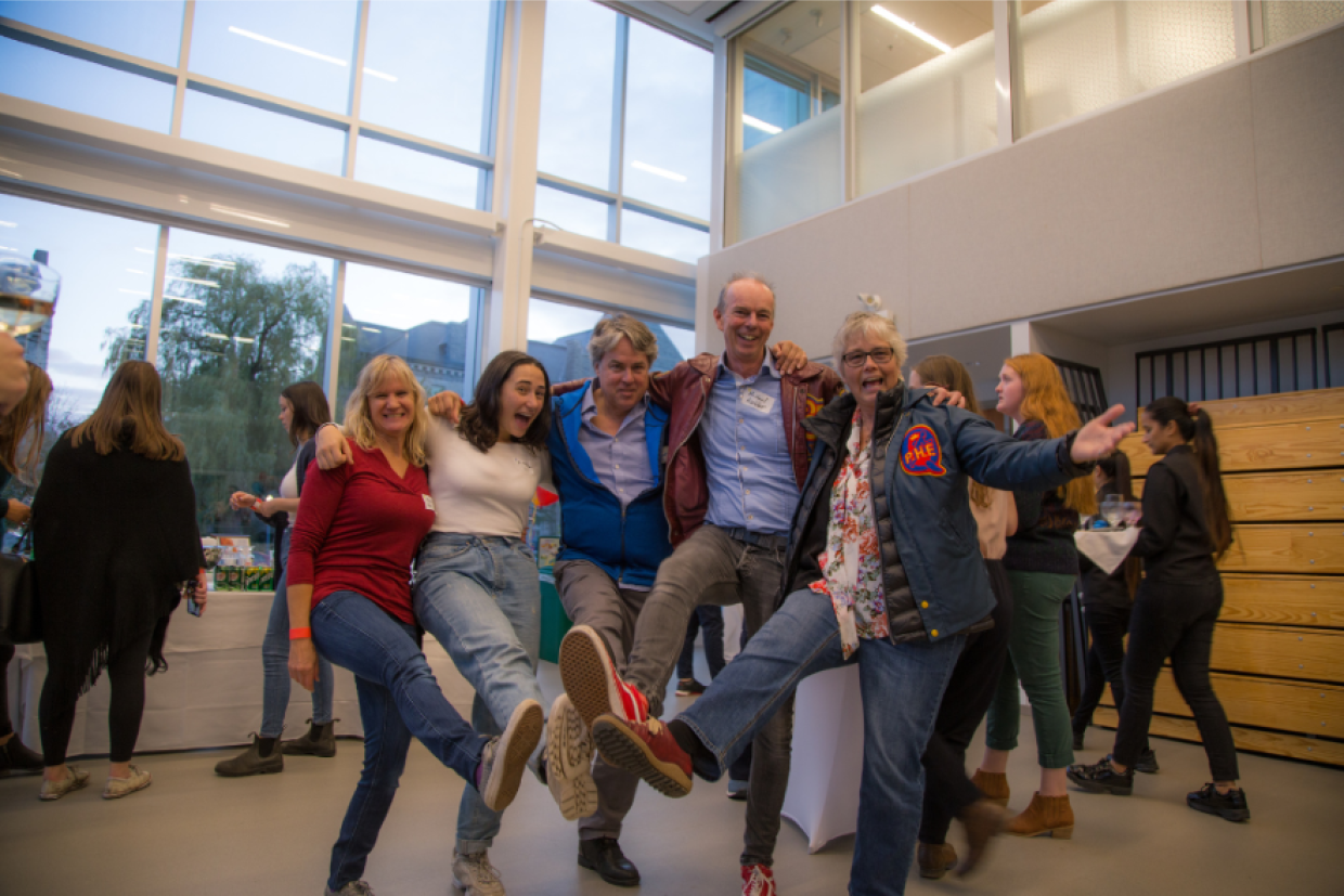 5 people with their arms connected over their shoulders doing the can can while smiling and laughing 