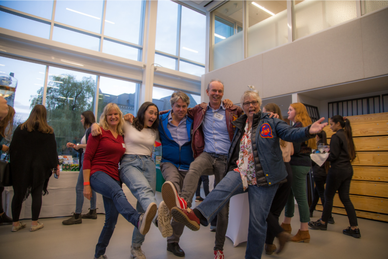 5 people with their arms connected over their shoulders doing the can can while smiling and laughing 