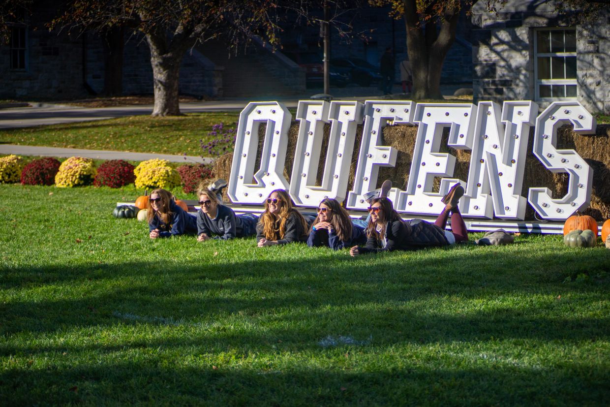 Several alumni lying on their bellies in front of a large, white sign that reads "Queen's"