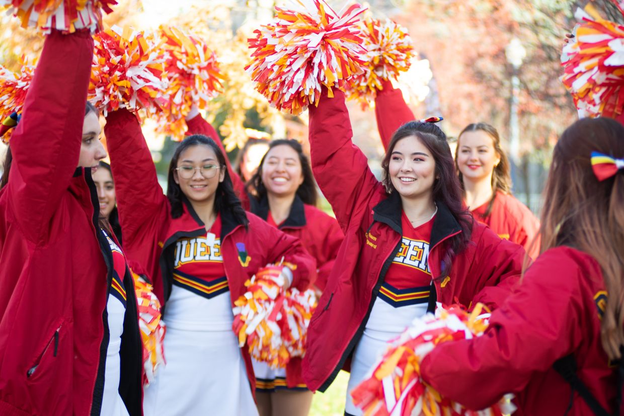 Cheerleaders standing on campus waving their pompoms, smiling.