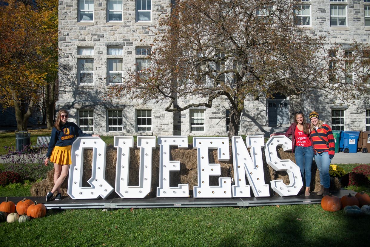 Alumni smiling while leaning on a light up sign that says "Queen's".